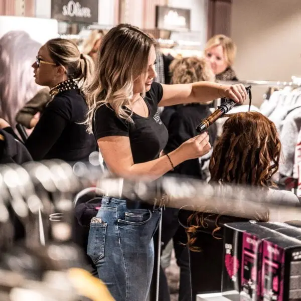 Pureday I en travl salon krøller en frisør med langt blondt hår kunstfærdigt en kundes brune lokker. Omgivet af livlig snak og forskellige salonprodukter udstråler atmosfæren naturlig skønhed og velvære, fokuseret på at forbedre hver enkelt kundes velvære gennem ekspert frisør.